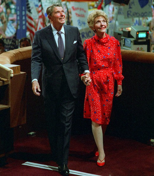 Ein Mann in schwarzer Jacke und weißem Hemd geht Hand in Hand mit einer Frau in rotem Kleid über eine rote Bühne, mit Menschen, Flaggen und Bannern im Hintergrund.